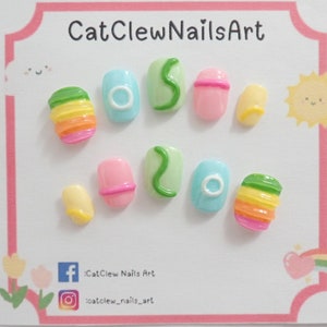 Peut inclure: Ensemble de dix faux ongles colorés avec différents motifs, notamment des rayures arc-en-ciel, des tourbillons verts et un ongle bleu avec un cercle blanc. Les ongles sont présentés sur un fond blanc avec un cadre rose.