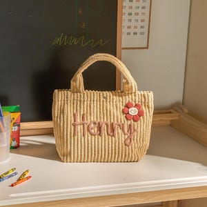 May include: A beige corduroy tote bag with the name "Henry" embroidered in pink thread. A red flower with a white center is attached to the bag. The bag has a handle and is sitting on a white surface.