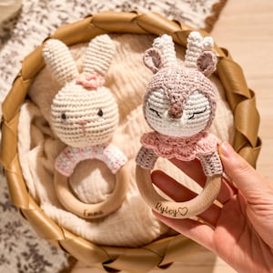 May include: Two handmade crochet baby rattles in a basket. One is a bunny with a pink bow, the other a deer with closed eyes. Both have wooden rings with names engraved. The bunny rattle is labeled "Emma", and the deer rattle is labeled "Ryley".