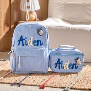 May include: A light blue corduroy backpack and lunch bag set. The name "Aiden" is embroidered in blue and white on both items, with an owl motif. The backpack has a front pocket and a top handle.