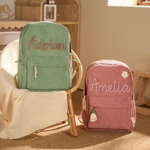 May include: Two corduroy backpacks, one light green with "Adrian" embroidered, and one pink with "Amelia". The pink backpack has a flower and a bell charm. Both have front pockets. They are on a chair, in a room.