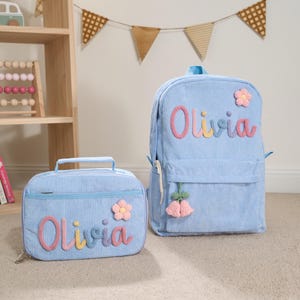 May include: A light blue corduroy backpack and matching lunch bag. Both items feature the name "Olivia" in colorful, embroidered letters and are decorated with flower accents. The backpack has a hanging tassel.