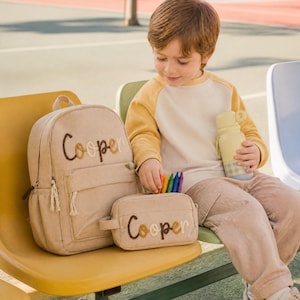 May include: A beige corduroy backpack and pencil case set, both embroidered with the name "Cooper". The backpack has a front pocket and a zip. A child holds a yellow water bottle and crayons. The set is designed for school.