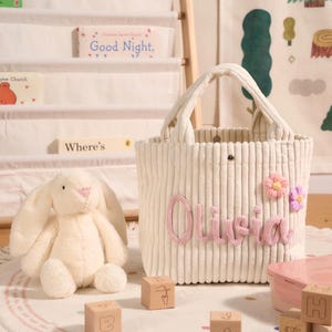May include: Cream-colored corduroy tote bag with the name "Olivia" in pink script, adorned with pink and purple fabric flowers. A plush white bunny and wooden blocks are nearby. The bag has a handle and is designed for carrying items.