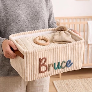 Peut inclure: Un panier de rangement rectangulaire en velours côtelé crème avec le prénom Bruce brodé en lettres de laine multicolores sur le devant. À l'intérieur se trouvent une couverture en tricot torsadé crème, un anneau de dentition et un bonnet.