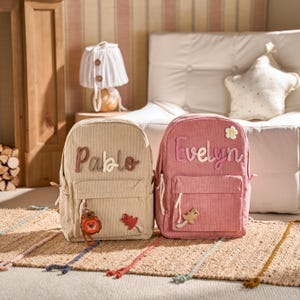 May include: Two corduroy backpacks, one beige and one pink, are shown. The beige backpack has the name "Pablo" stitched on it, and the pink backpack has the name "Evelyn". Both have front pockets and decorative charms.