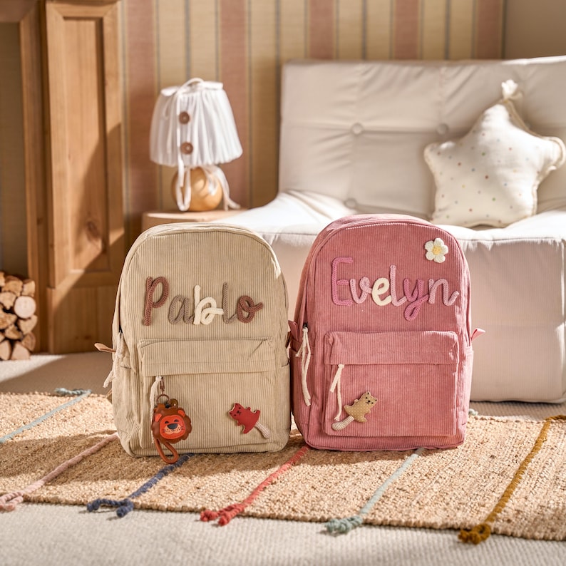 May include: Two corduroy backpacks, one beige and one pink, are shown. The beige backpack has the name "Pablo" stitched on it, and the pink backpack has the name "Evelyn". Both have front pockets and decorative charms.