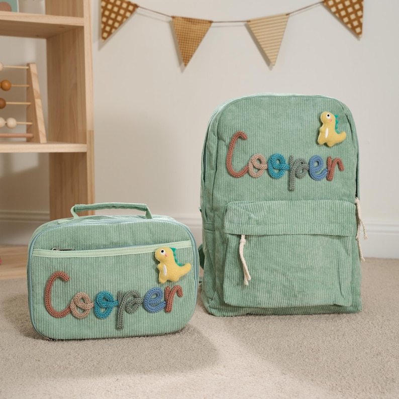 Puede incluir: Un juego a juego de mochila y bolsa de almuerzo de pana verde menta. El nombre "Cooper" est&aacute; cosido en cada art&iacute;culo con hilo colorido. Cada bolsa tiene una aplicaci&oacute;n de dinosaurio amarillo.