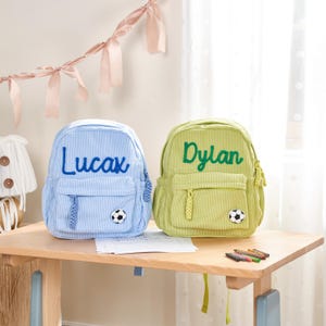 May include: Two corduroy backpacks, one light blue with "Lucax" embroidered in blue, and one light green with "Dylan" embroidered in green. Each backpack has a soccer ball patch and a front pocket.