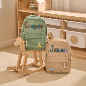 May include: Two corduroy backpacks are shown, one green and one tan. The green backpack has the name "Cooper" in colourful letters, and a yellow dinosaur patch. The tan backpack has the name "Jaxon" in blue letters, and a blue dinosaur patch.