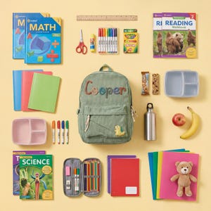 May include: A collection of school supplies, including books, notebooks, pens, pencils, crayons, scissors, glue, a backpack with the name "Cooper", a lunchbox, a water bottle, a red apple, and a banana. A teddy bear is also included.