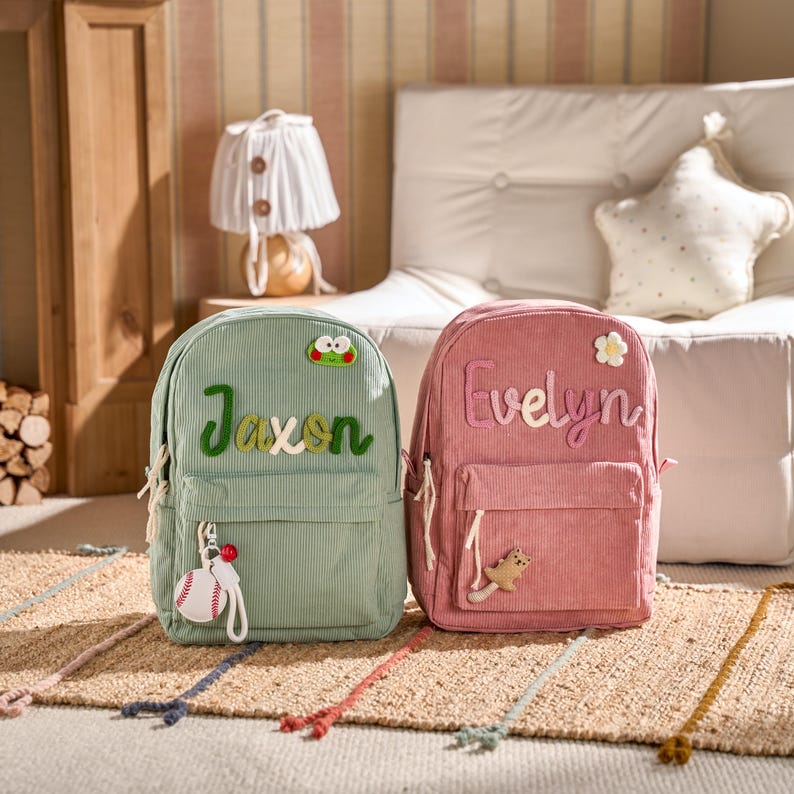 May include: Two corduroy backpacks, one light green and one pink, are displayed. The green backpack has the name "Jaxon" embroidered on it, along with a baseball keychain. The pink backpack is embroidered with the name "Evelyn" and a flower.