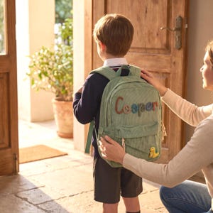 May include: A sage green corduroy backpack with the name "Cooper" embroidered in colourful letters. The backpack features a small yellow dinosaur patch. A person is adjusting the backpack on a child wearing a navy blue sweater and shorts.