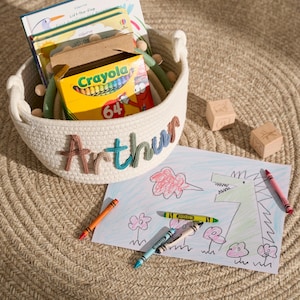 May include: A white rope basket with the name "Arthur" embroidered on the side. The basket is filled with children's books and a box of crayons. A drawing of a dragon blowing fire is on a white sheet of paper with crayons next to it.