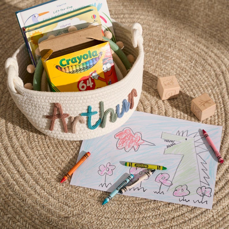 May include: A white rope basket with the name "Arthur" embroidered on the side. The basket is filled with children's books and a box of crayons. A drawing of a dragon blowing fire is on a white sheet of paper with crayons next to it.
