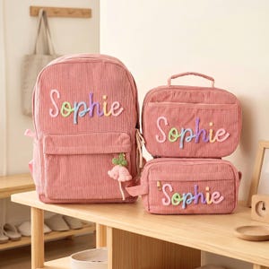 May include: A set of three dusty pink corduroy bags including a large backpack, a rectangular lunch bag, and a smaller pencil case. Each bag features the name Sophie embroidered in a colourful, multi-toned gradient yarn and a small flower charm.