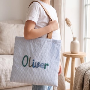 Peut inclure: Un sac fourre-tout en seersucker bleu clair et blanc avec le prénom Oliver brodé en lettres cursives dans des tons de bleu et de vert. Le sac possède une bandoulière en tissu beige et une forme rectangulaire avec une finition texturée.