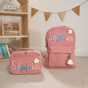Puede incluir: Mochila y bolsa de almuerzo a juego de pana rosa. Ambos art&iacute;culos presentan el nombre "Sophia" en letras bordadas de colores y una aplicaci&oacute;n de flor blanca. La mochila tiene una borla y la bolsa de almuerzo un asa.