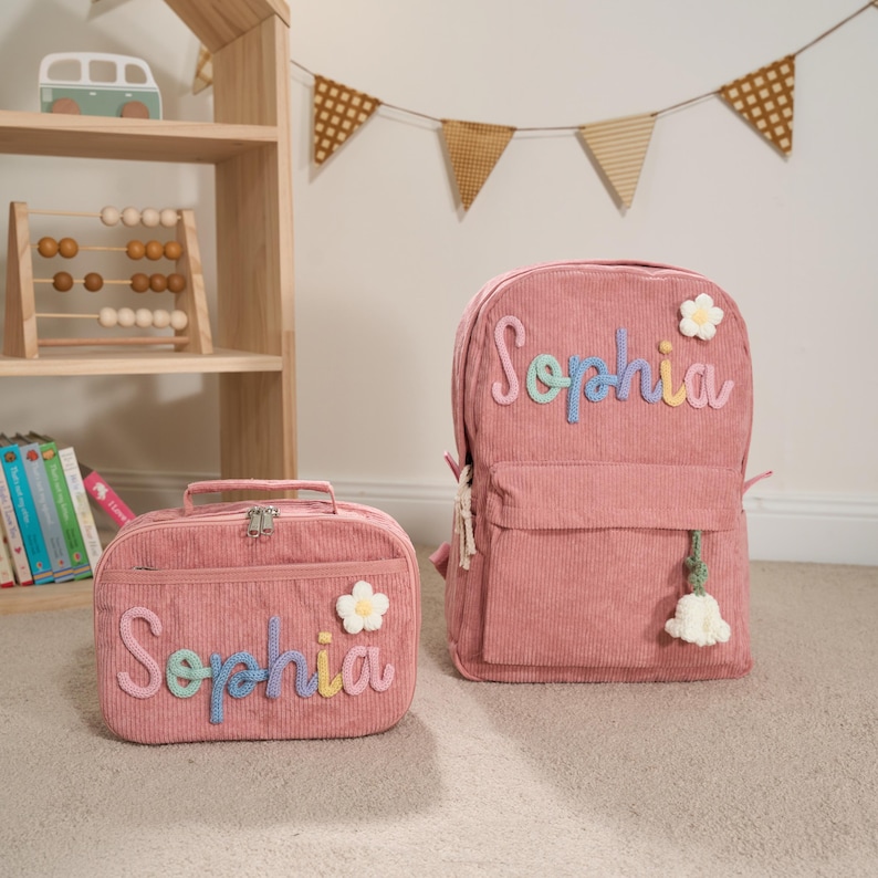 Puede incluir: Mochila y bolsa de almuerzo a juego de pana rosa. Ambos art&iacute;culos presentan el nombre "Sophia" en letras bordadas de colores y una aplicaci&oacute;n de flor blanca. La mochila tiene una borla y la bolsa de almuerzo un asa.