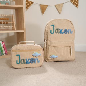 Puede incluir: Juego de mochila y bolsa de almuerzo de pana beige. Ambos art&iacute;culos presentan el nombre "Jaxon" en escritura azul y verde, junto con una aplicaci&oacute;n de dinosaurio azul claro. La mochila tiene un bolsillo frontal y correas ajustables.