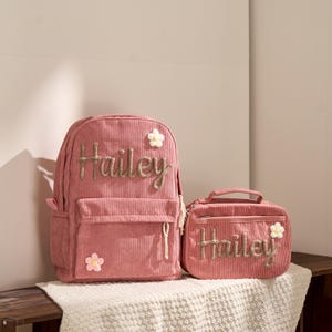 May include: Pink corduroy backpack and matching lunch bag set. Both items feature the name "Hailey" embroidered in gold thread, along with floral embellishments. The backpack has a front pocket and side pocket.