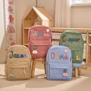 May include: Four corduroy backpacks in tan, pink, blue, and green, each with a name embroidered on the front. The backpacks have a front pocket, a top handle, and decorative appliqu&eacute;s like flowers and a dinosaur.
