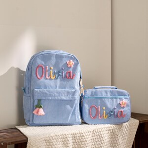 Peut inclure: Un sac à dos et un sac à déjeuner assortis en velours côtelé bleu clair. Les deux articles présentent le nom "Olivia" en lettres brodées colorées et sont décorés d'accents floraux. Le sac à dos a une poche avant et un pompon rose.