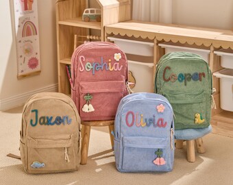 May include: Four corduroy backpacks in tan, pink, blue, and green, each with a name embroidered on the front. The backpacks have a front pocket and decorative appliques, including flowers and a dinosaur.
