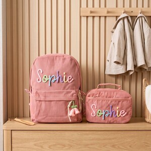 May include: A dusty pink corduroy backpack and a matching lunch bag, both featuring the name Sophie in colourful, raised, cursive lettering. The backpack includes a small, dangling pink flower charm attached to the front zipper pull.