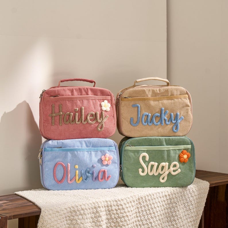 May include: Four corduroy lunch bags in pink, tan, blue, and green. Each bag has a different name embroidered on it: Hailey, Jacky, Olivia, and Sage. Each bag has a flower accent. The bags have a zipper closure and a handle.