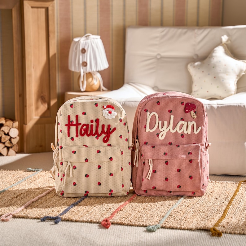 May include: Two corduroy backpacks, one beige and one pink, each with a name embroidered on the front. The beige backpack has the name "Haily" in red script and a Santa Claus patch. The pink backpack has the name "Dylan" in white script and a mushroom patch.