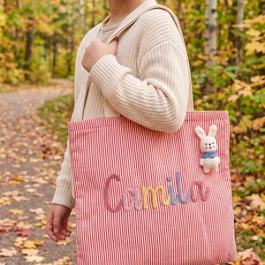 Peut inclure: Un sac fourre-tout à rayures rouges et blanches portant le prénom Camila brodé en laine multicolore sur le devant. Une petite figurine de lapin blanc en peluche avec un col à motifs bleus est fixée dans le coin supérieur droit du sac.