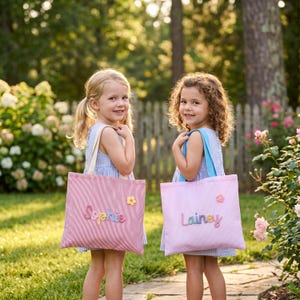 Peut inclure: Deux sacs fourre-tout rectangulaires avec des anses en tissu. Le sac de gauche est à rayures rouges et blanches avec le prénom Sophie en lettres colorées et une petite fleur jaune. Le sac de droite est rose clair avec le prénom Lainey en lettres colorées et une petite fleur rose.