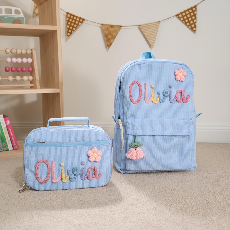 May include: A light blue corduroy backpack and matching lunch bag. Both items feature the name "Olivia" in colorful, embroidered letters and are decorated with flower accents. The backpack has a hanging tassel.