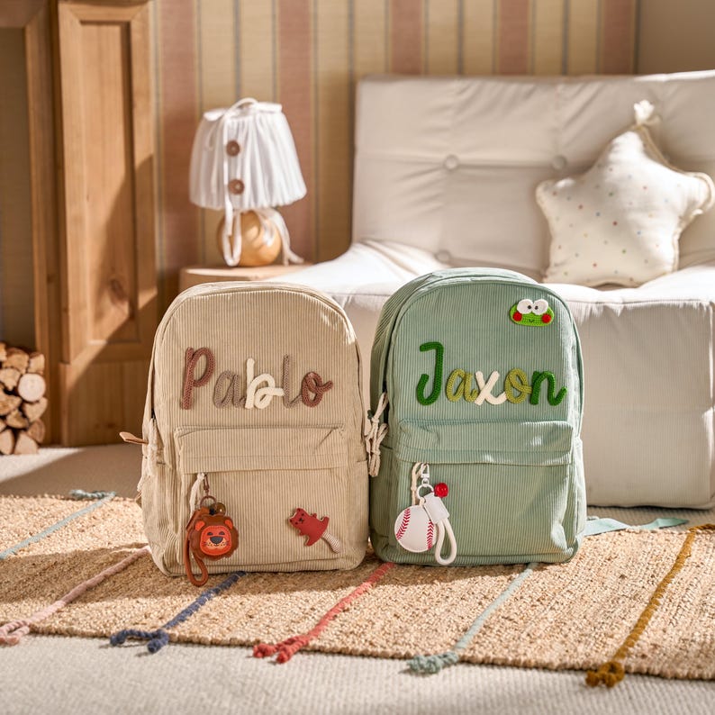 May include: Two corduroy backpacks, one beige and one sage green, are displayed on a woven rug. The beige backpack has the name "Pablo" and a lion keychain. The green backpack has the name "Jaxson" and a baseball keychain.
