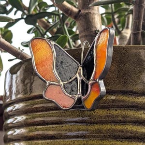 May include: A stained glass butterfly with orange, black and pink wings. The butterfly is attached to a brown ceramic pot.