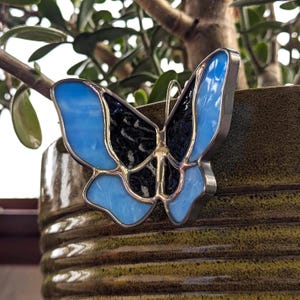 May include: A stained glass butterfly ornament with blue and black wings. The butterfly is hanging from a plant pot.