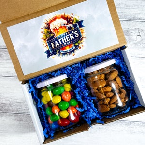 May include: A brown cardboard box with a blue and white Father's Day themed lid. The box contains two glass jars, one filled with colorful candies and the other with almonds.