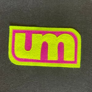 May include: A felt patch with the letters "um" in pink on a bright yellow background.