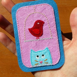 May include: A felt applique with a blue border, pink background, and embroidered images of a red bird in a cage and a blue cat.