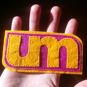 May include: A felt patch with the letters "UM" in yellow with pink stitching on a pink background.
