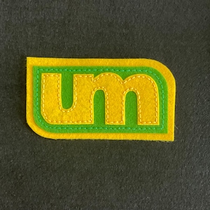 May include: A yellow felt patch with green stitching, shaped like a rounded rectangle, with the letters "UM" in green stitching on a yellow background.