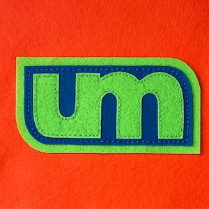 May include: A green felt patch with the letters "UM" in blue felt, outlined in blue stitching.