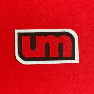 May include: A white patch with a black border and red stitching. The letters "UM" are stitched in red on the patch.