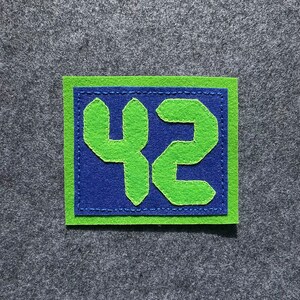 May include: A green felt patch with the number 42 stitched on it in a green thread. The number is on a blue felt background and the patch is surrounded by a green felt border.