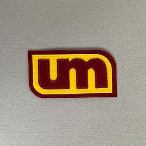 May include: A maroon felt patch with yellow stitching and the letters "UM" in a bold font.