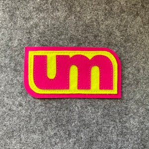 May include: A pink felt patch with the letters "UM" in yellow felt, outlined in pink stitching.