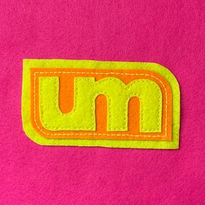 May include: A felt patch with the letters "um" in a lime green color with a bright orange outline. The patch is sewn with a white thread.