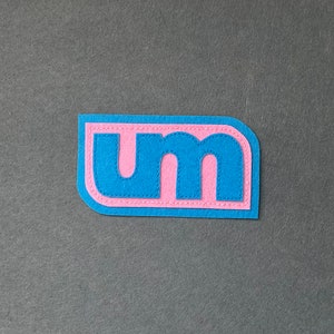 May include: A blue felt patch with the letters "UM" in pink felt, stitched with pink thread.