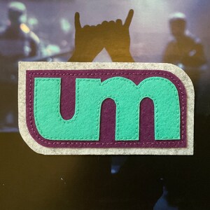 May include: A felt patch with the letters "um" in a teal green color with a purple outline. The patch is surrounded by a white border with a stitched edge.